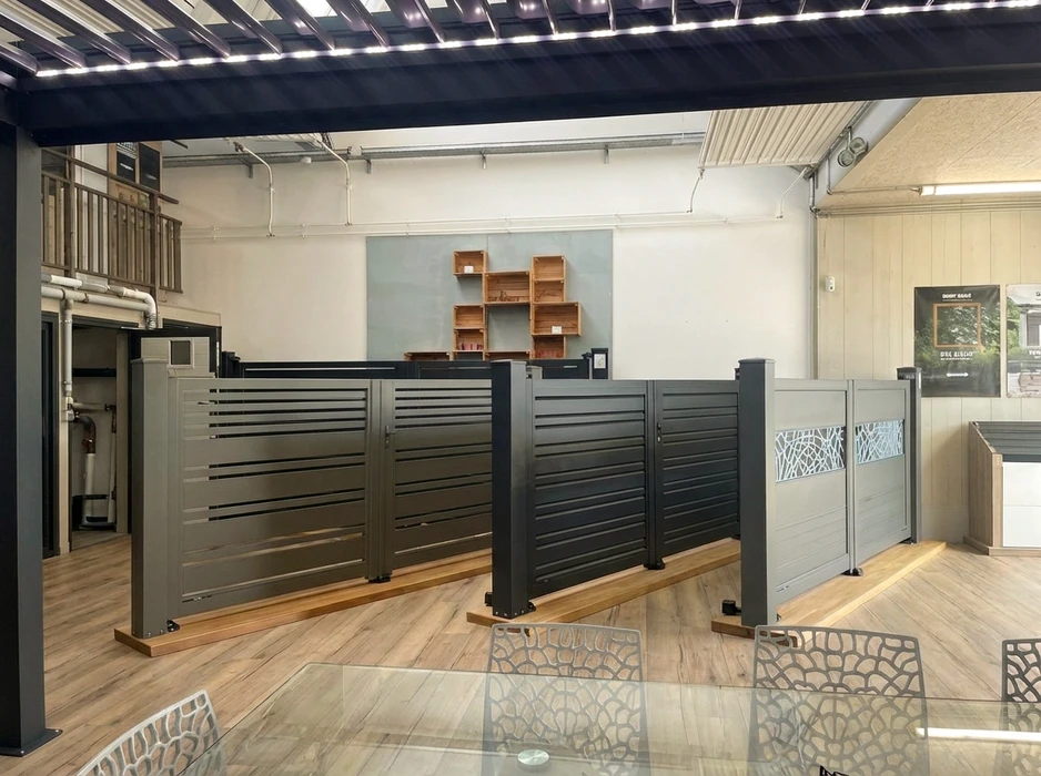 Intérieur d'un showroom de menuiserie extérieure bien éclairé, avec exposition de plusieurs produits : portails (en aluminium et fer forgé), pergolas bioclimatiques, stores banne, échantillons de fenêtres et portes affichant différentes finitions (couleurs, matériaux). Un couple ou deux clients sont visibles en arrière-plan, touchant et examinant les matériaux, discutant avec un vendeur souriantes devant des modules de démonstration. L'ambiance est accueillante, le showroom spacieux et bien rangé, mettant en avant la qualité et la variété des solutions proposées.